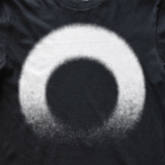 A close-up of a black modular synth t-shirt featuring a large, circular graphic print with a halftone texture. The design consists of a solid black center surrounded by a white ring that fades into black at the bottom edge.