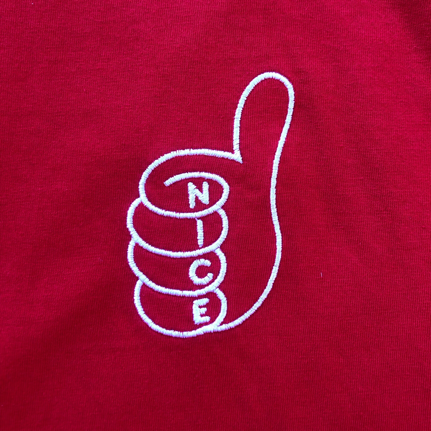 A close-up of a red fabric surface featuring a white embroidered outline of a thumbs-up hand with the letters 'N', 'I', 'C', 'E' stacked vertically inside the fingers.