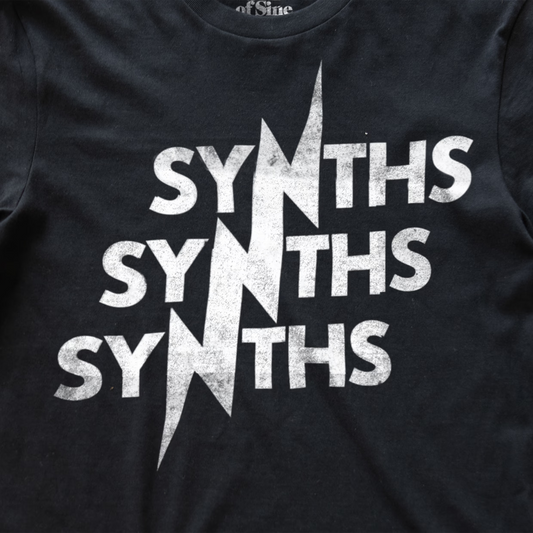 A close-up of a black synth inspired t-shirt featuring a distressed white graphic print that reads 'SYNTHS SYNTHS SYNTHS' with a jagged lightning bolt design running through the middle.