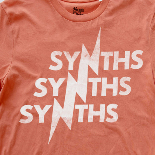 A close-up of a coral pink synth inspired t-shirt featuring a distressed white graphic print that reads 'SYNTHS' repeated vertically with a lightning bolt motif running through the center.