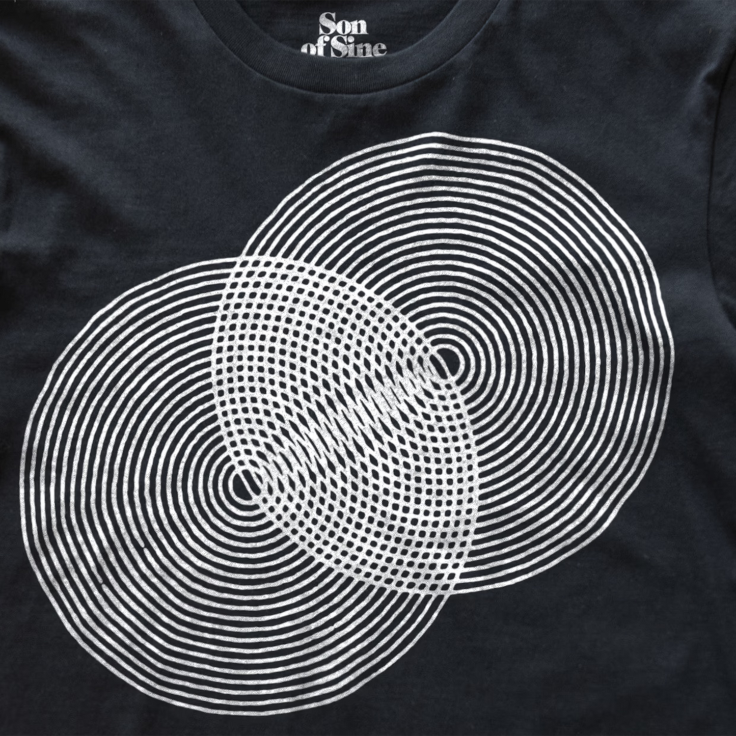 A close-up of a black modular synth t-shirt featuring a white graphic print of two overlapping circles made of concentric rings and dots. The inner collar shows the text 'Son of Sine' in white.