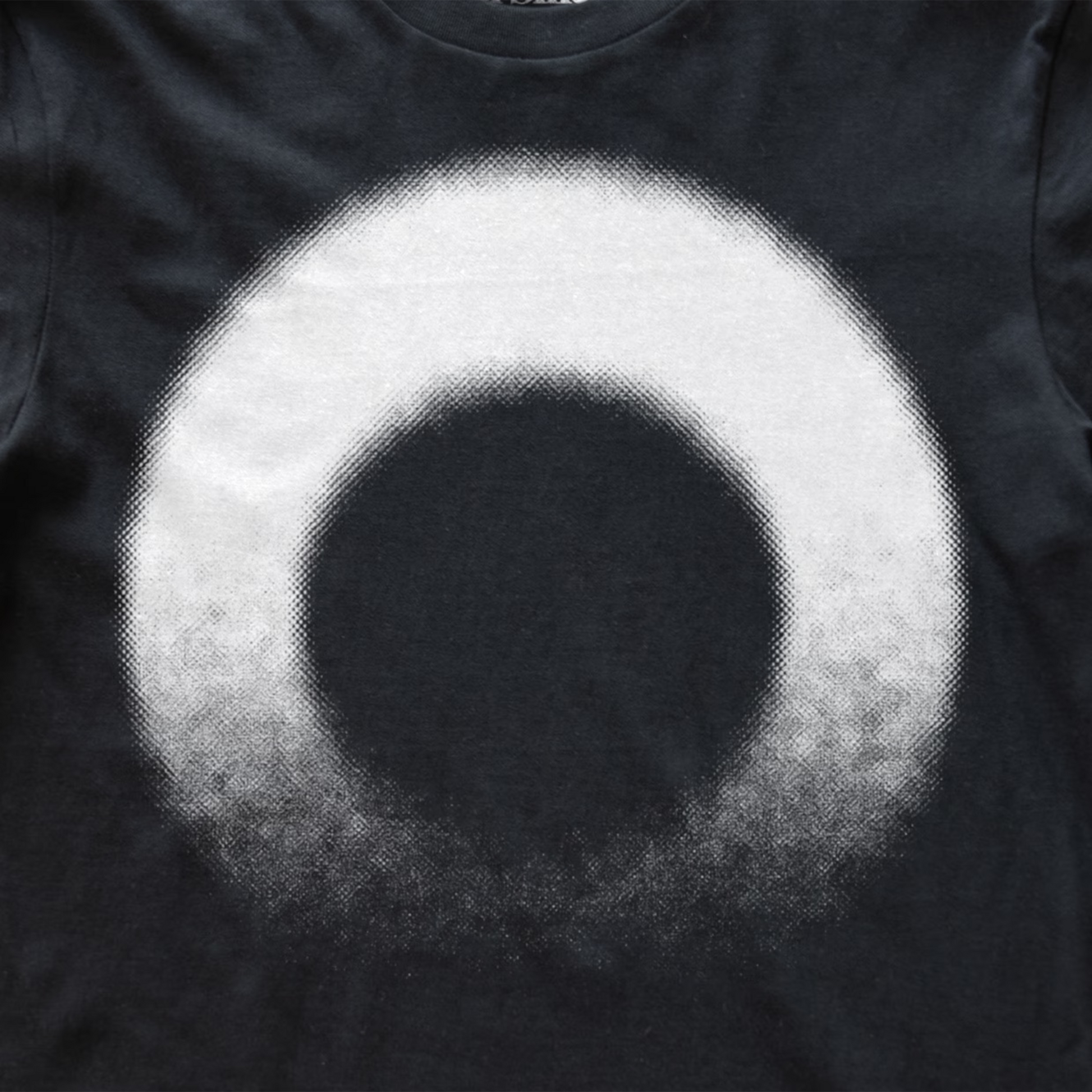 A close-up of a black modular synth t-shirt featuring a large, circular graphic print with a halftone texture. The design consists of a solid black center surrounded by a white ring that fades into black at the bottom edge.