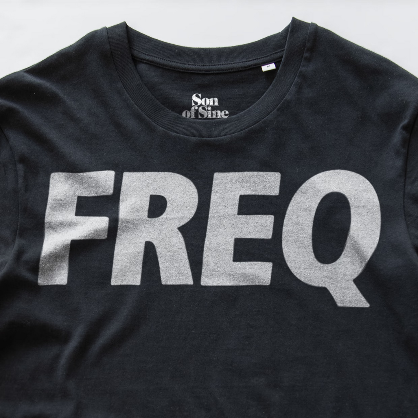 A close-up of a black electronic music club wear t-shirt featuring the word 'FREQ' printed across the chest in a large, ALL CAPS white font. The inner collar displays the brand name 'Son of Sine'.