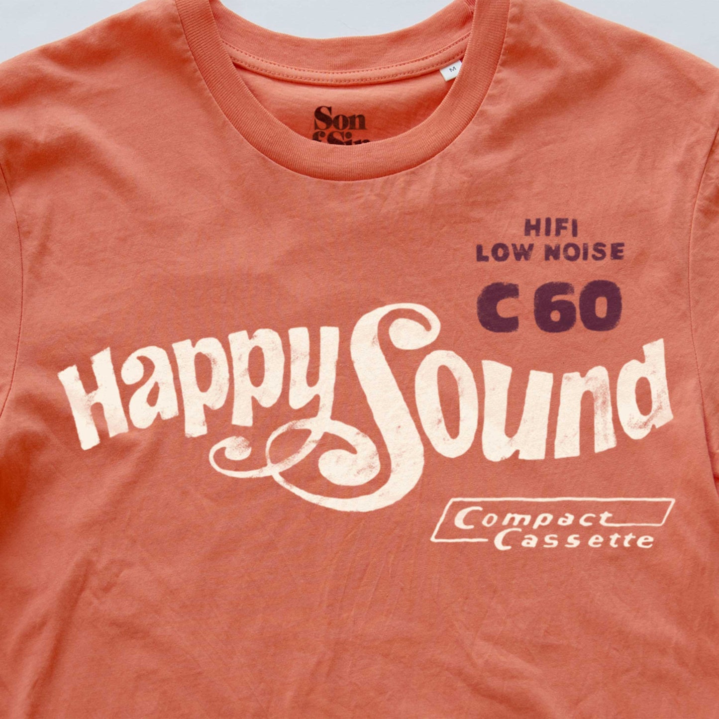 A close-up of a coral coloured synth inspired t-shirt featuring a vintage-style cassette sleeve graphic print that reads 'Happy Sound C60 HIFI LOW NOISE' and 'Compact Cassette'. 