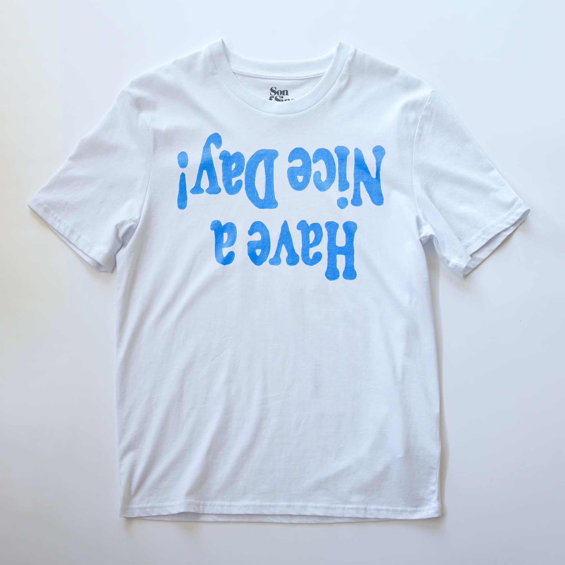 A white electronic music festival t-shirt with the phrase 'Have a Nice Day!' printed upside down in blue bubble letters on the front.