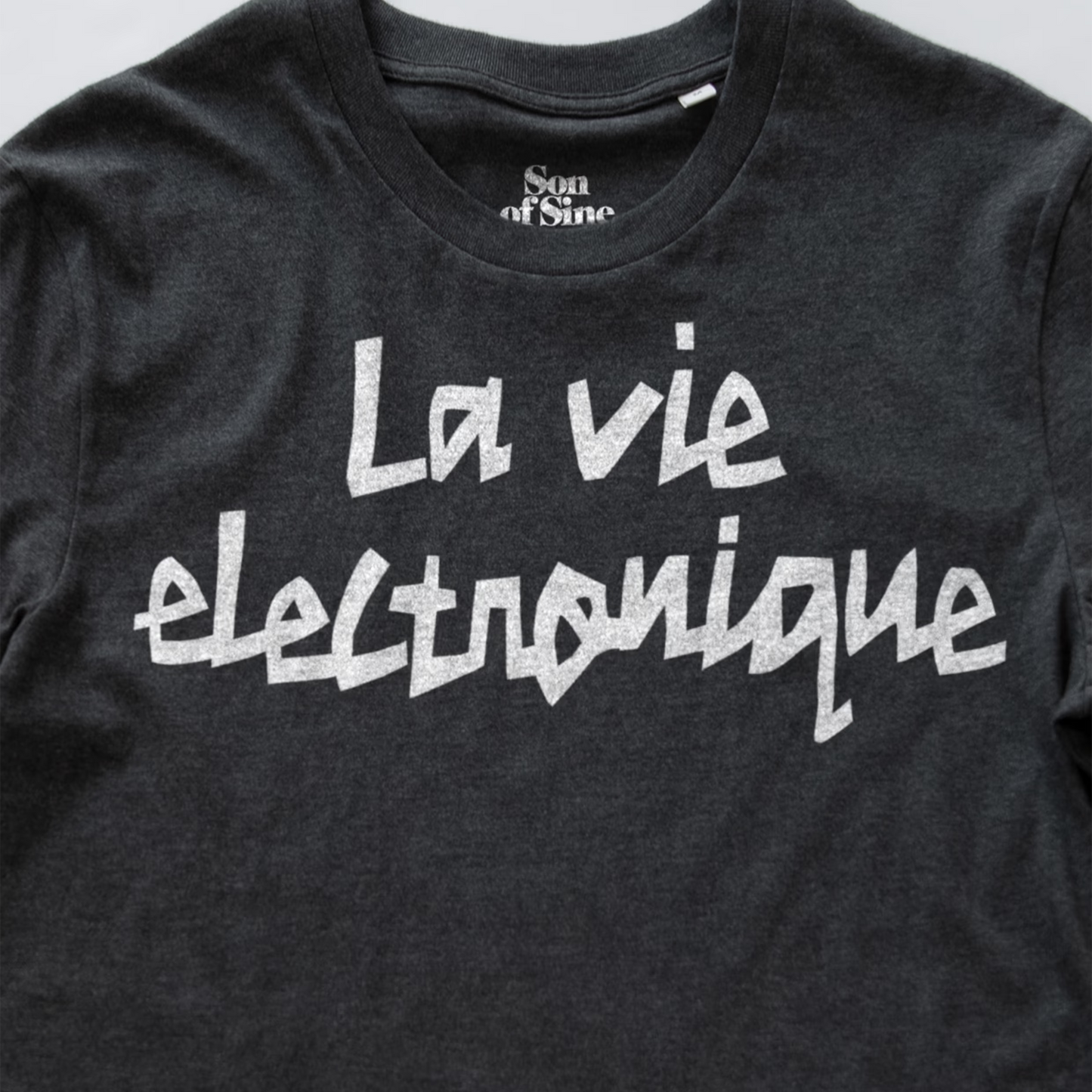 A close-up of a dark heather gray synth t-shirt featuring the phrase 'La vie electronique' printed across the chest in a white, stylized handwritten font. The inner collar label shows the brand 'Son of Sine'.
