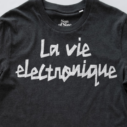 A close-up of a dark heather gray synth t-shirt featuring the phrase 'La vie electronique' printed across the chest in a white, stylized handwritten font. The inner collar label shows the brand 'Son of Sine'.