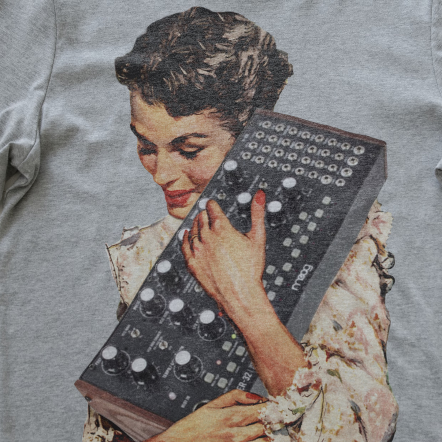 A close-up of a light gray heather synth t-shirt featuring a vintage-style graphic print of a woman holding a mother 32 modular synthesizer.