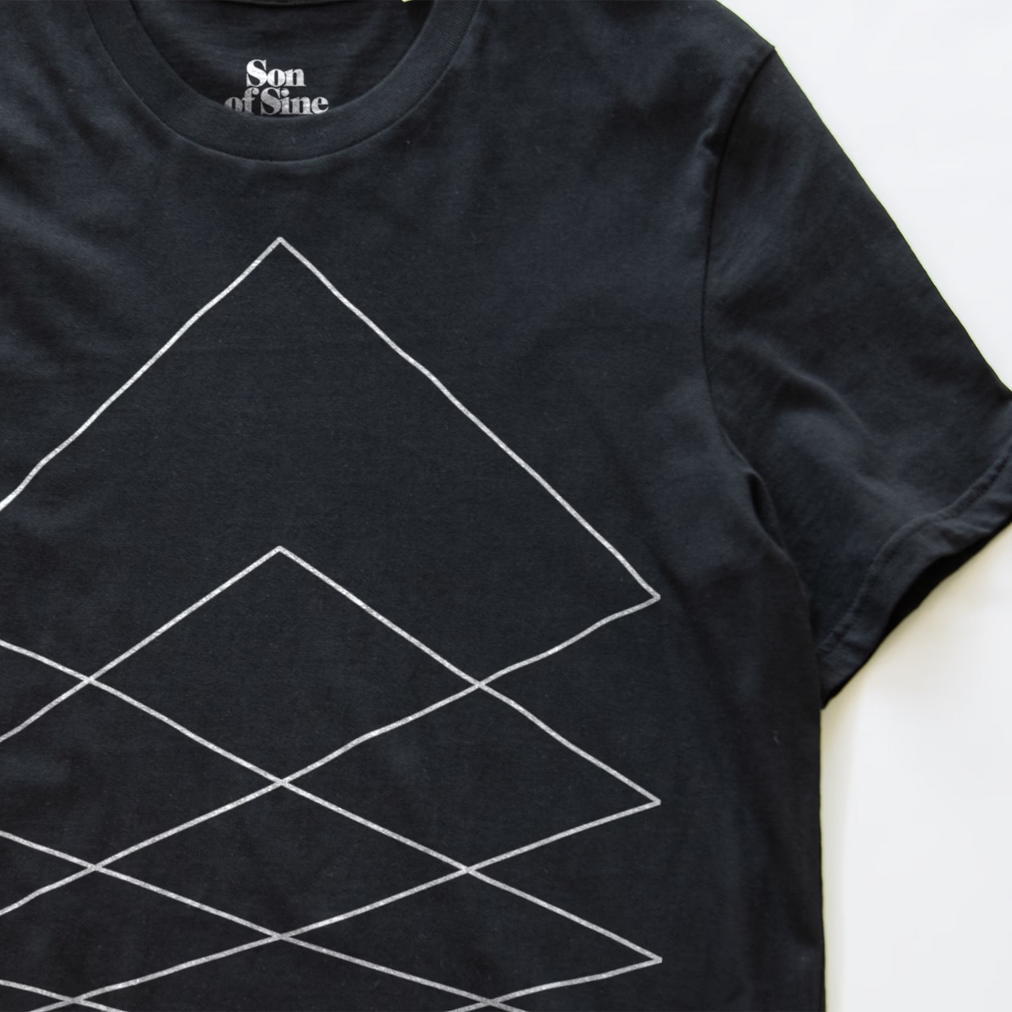 A close-up of a black modular synth t-shirt featuring a white geometric line pattern of nested diamonds, with the 'Son of Sine' brand logo printed inside the collar.