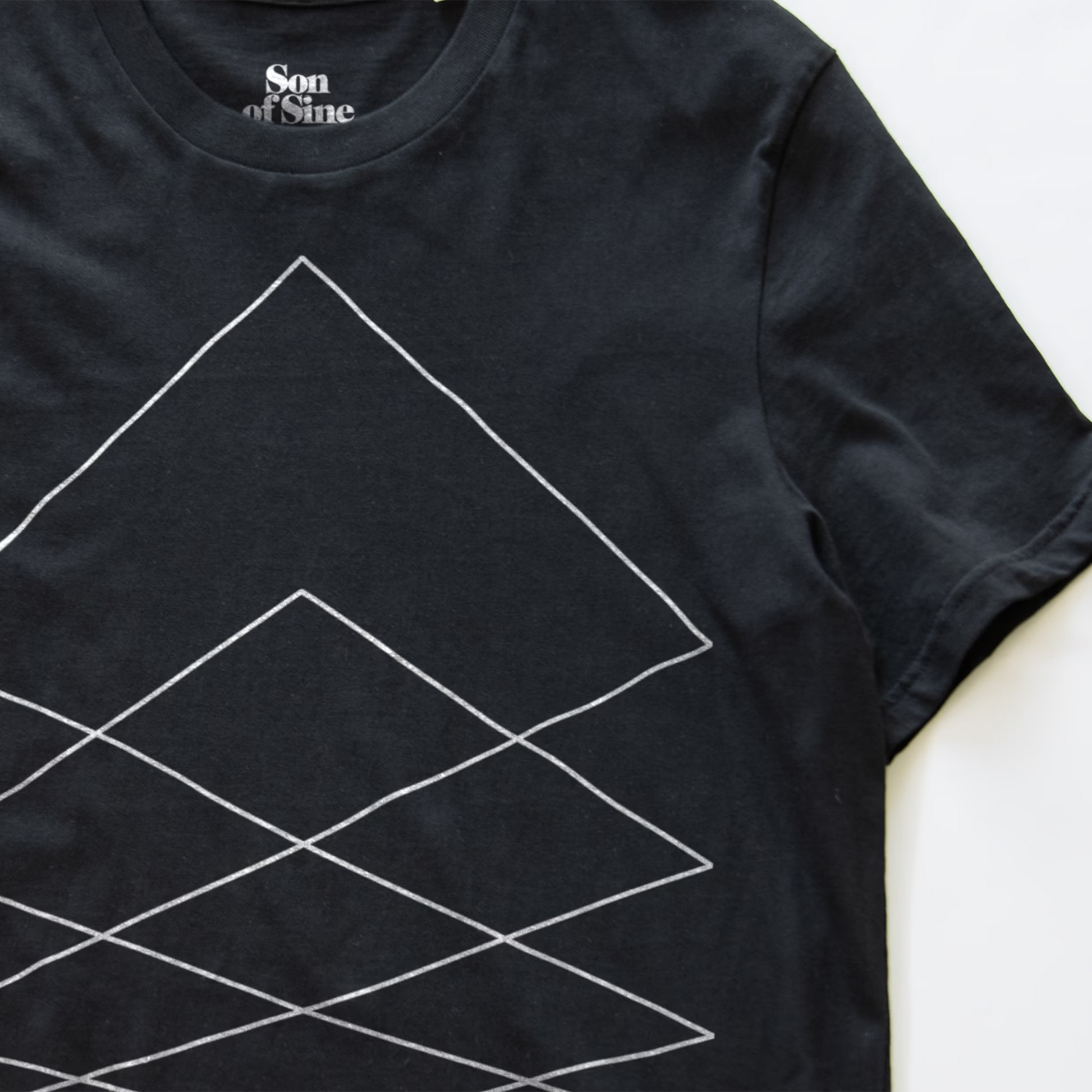 A close-up of a black modular synth t-shirt featuring a white geometric line pattern of nested diamonds, with the 'Son of Sine' brand logo printed inside the collar.