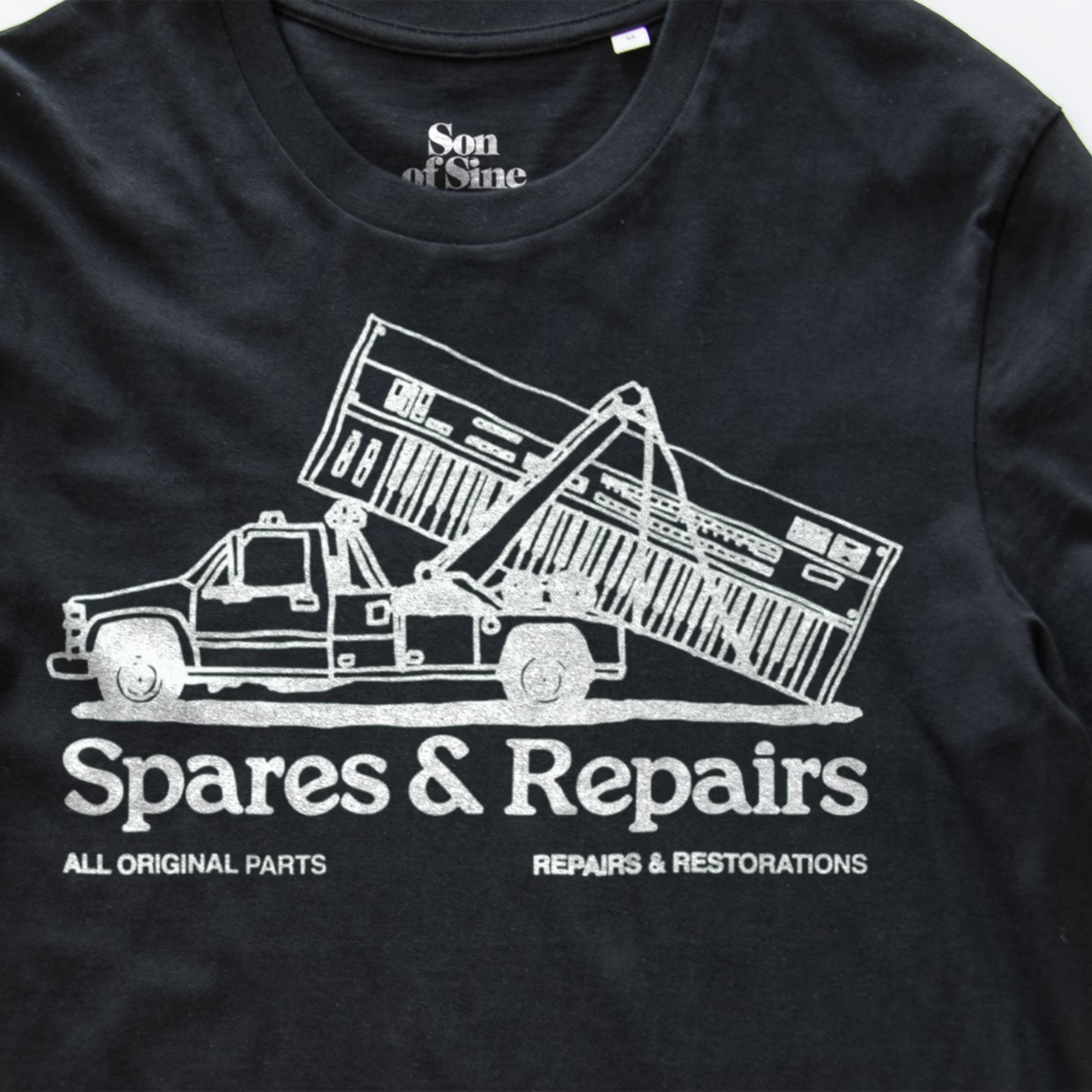 A close-up of a black electronic music t-shirt featuring a white graphic of a tow truck lifting a synth with the text 'Spares & Repairs' and 'All Original Parts Repairs & Restorations'. The inner collar shows the brand 'Son of Sine'.